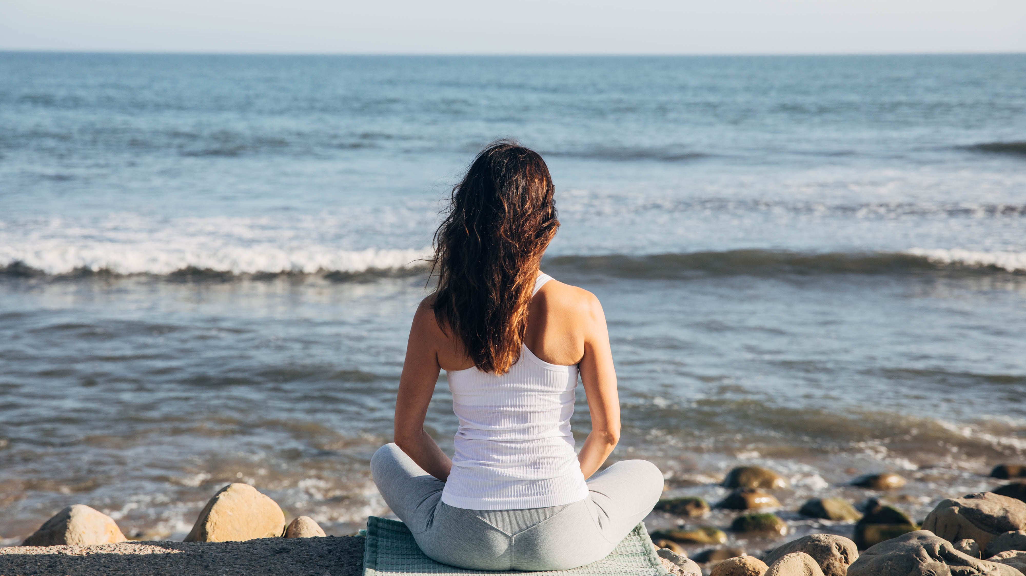 Woman in seated meditation on sage Ritual Rug looking at the ocean. Comfortable for any yoga or meditation practice, The Ritual Rug offers cushion and support.