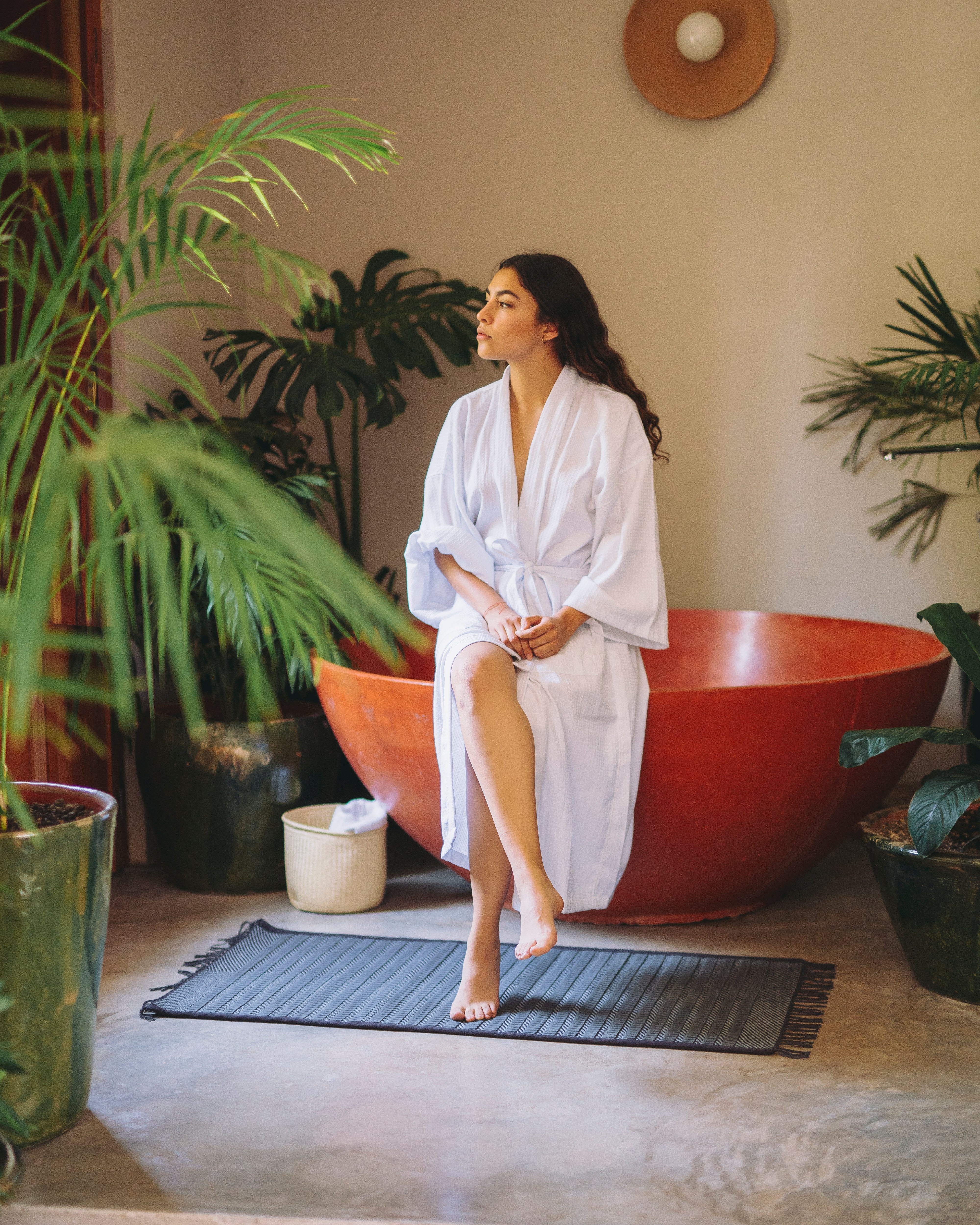Woman in a white robe sitting on a large red bowl surrounded by plants in a room with a neutral color palette.