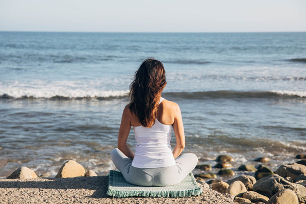 Woman in seated meditation on sage Ritual Rug looking at the ocean. Comfortable for any yoga or meditation practice, The Ritual Rug offers cushion and support.